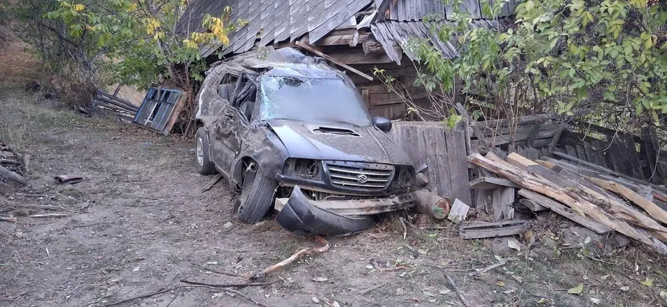 Un autoturism de culoare închisă, avariat, este poziționat lângă o construcție din lemn și stive de scânduri într-o zonă cu vegetație.