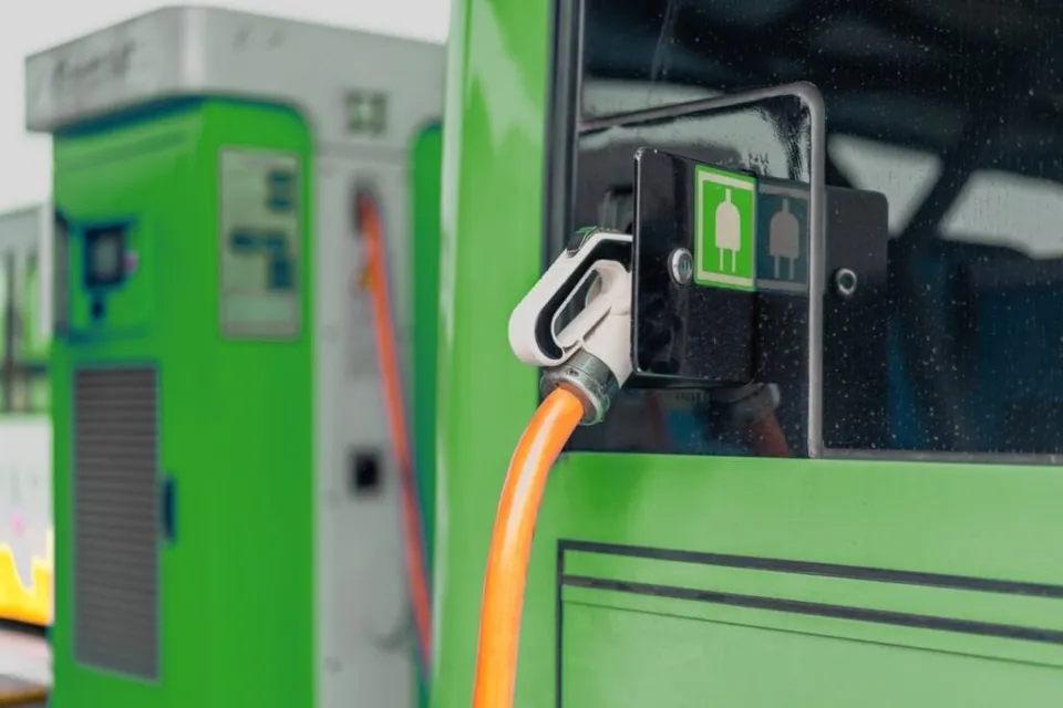 Un cablu portocaliu cu un conector alb este conectat la o stație de încărcare pe un vehicul de culoare verde.