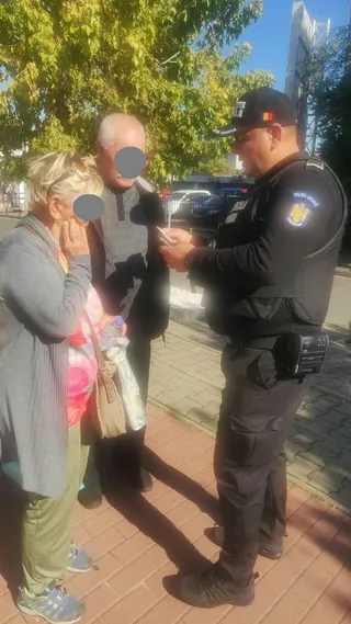 Un polițist în uniformă neagră stă pe un trotuar lângă o femeie și un bărbat sub copaci.