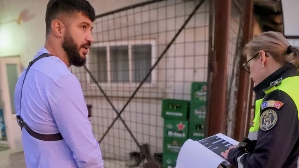 Un bărbat cu barbă și cămașă albastră stă în fața unei femei care poartă o uniformă neagră și galbenă cu un emblema pe braț.