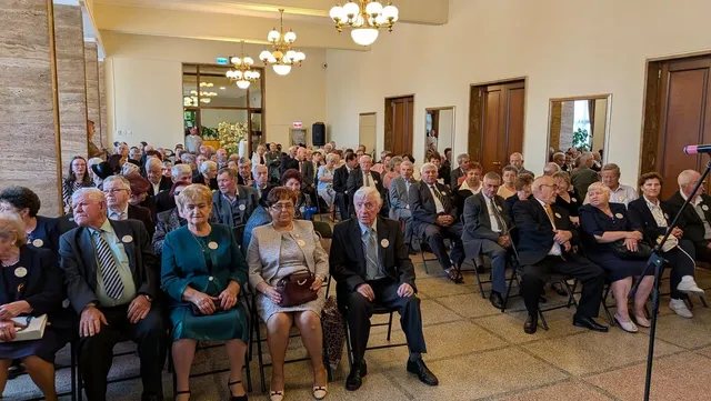 Un grup de persoane în vârstă stă așezat pe scaune într-o sală de evenimente cu lustre suspendate și uși din lemn.