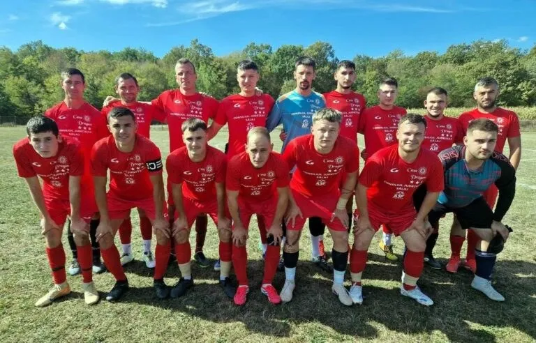 Un grup de fotbaliști în echipamente roșii și albastre pozează împreună pe un teren de fotbal cu copaci în fundal.