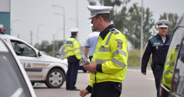 Un polițist în uniformă galbenă și neagră ține un dispozitiv în mână, în timp ce alți doi polițiști stau pe o stradă lângă mașini.