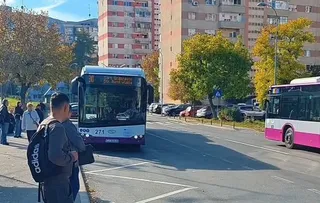 Un autobuz alb și albastru circulă pe un drum asfaltat lângă un autobuz roz și alb, în fața unor clădiri de locuințe și copaci.