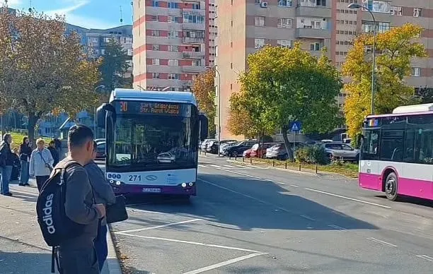 Un autobuz alb și albastru circulă pe un drum asfaltat lângă un autobuz roz și alb, în fața unor clădiri de locuințe și copaci.