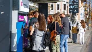 Un grup de tineri stă în picioare pe un trotuar lângă un automat de bilete și o stație de autobuz.