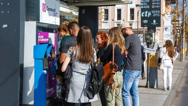 Un grup de tineri stă în picioare pe un trotuar lângă un automat de bilete și o stație de autobuz.