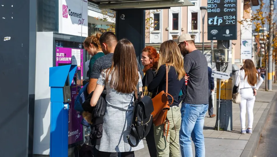 Un grup de tineri stă în picioare pe un trotuar lângă un automat de bilete și o stație de autobuz.