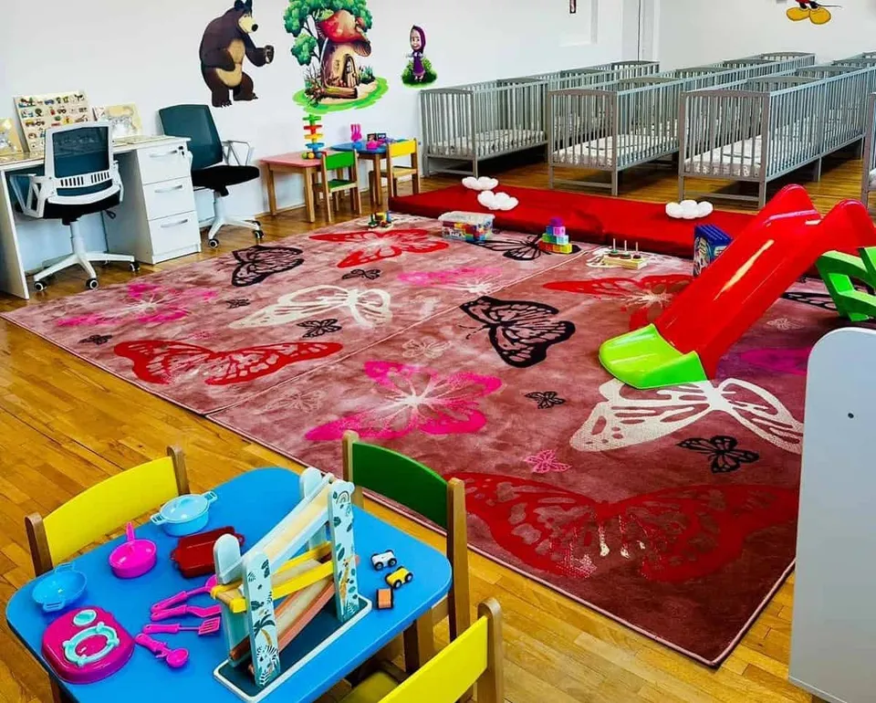 O cameră de joacă pentru copii conține un covor cu fluturi, un tobogan roșu, cots metalice și mese mici cu scaune colorate.