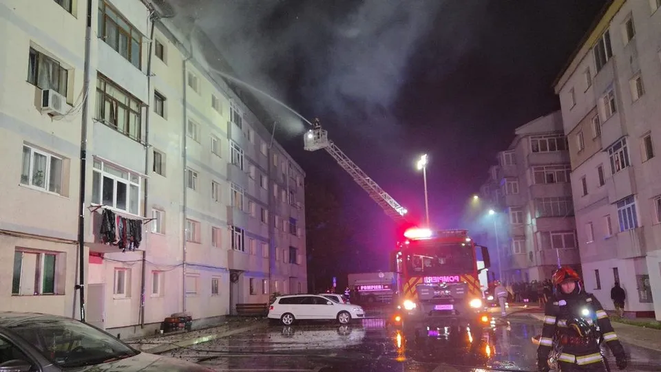 Un autospecial de pompieri cu lumină roșie este parcat pe un drum umed lângă un bloc, sub o scară de salvare extinsă spre acoperiș.