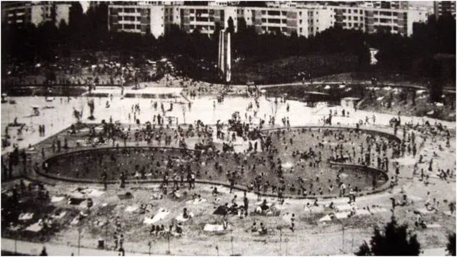 O mulțime de oameni se află într-o zonă deschisă lângă o piscină cu forme curbe, în fața unor blocuri de locuințe.