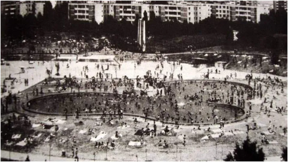 O mulțime de oameni se află într-o zonă deschisă lângă o piscină cu forme curbe, în fața unor blocuri de locuințe.