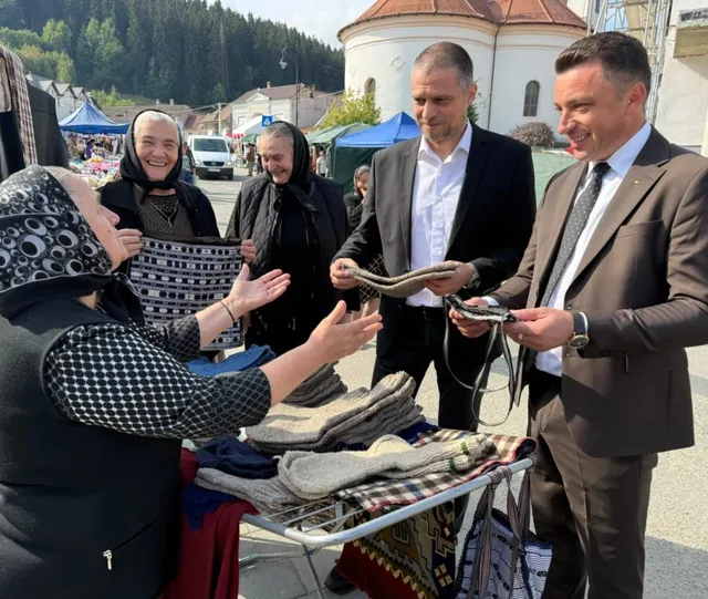 Un grup de persoane stă în fața unui stand cu obiecte din lână, în timp ce doi bărbați în costume privesc materialele textile.
