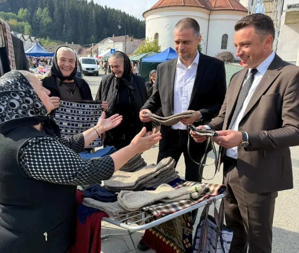 Un grup de persoane stă în fața unui stand cu obiecte din lână, în timp ce doi bărbați în costume privesc materialele textile.