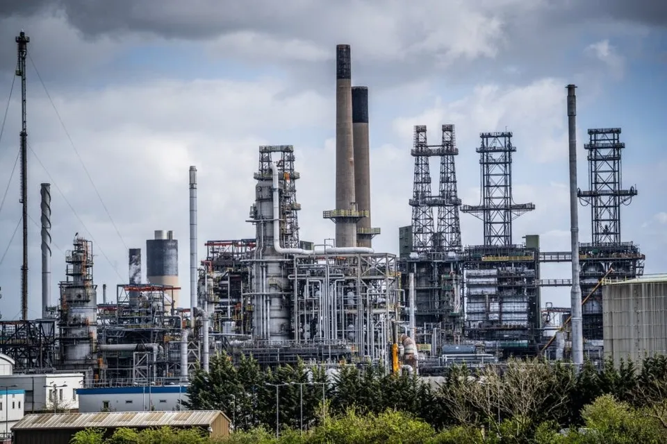 O fabrică industrială cu numeroase turnuri înalte, conducte metalice și hornuri sub un cer înnorat, situată în spatele unor copaci verzi.