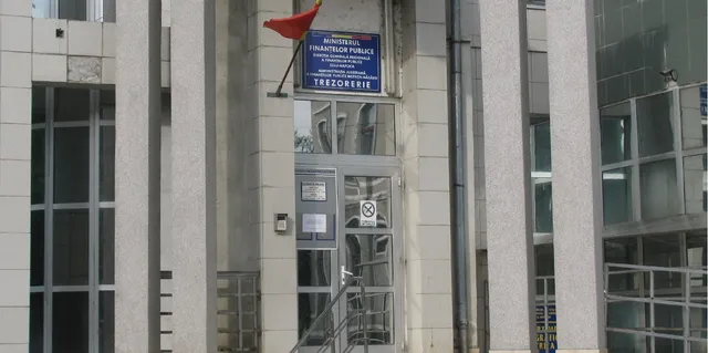 O clădire modernă cu fațadă din beton și sticlă prezintă o intrare cu uși de sticlă și un steag roșu pe o sârmă.