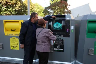 Un bărbat și o femeie stau lângă containere de colectare a deșeurilor, acesta arătând spre o deschidere de colectare.