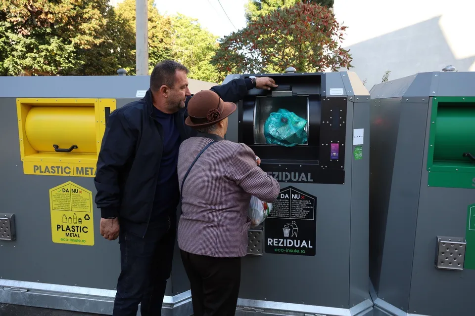 Un bărbat și o femeie stau lângă containere de colectare a deșeurilor, acesta arătând spre o deschidere de colectare.