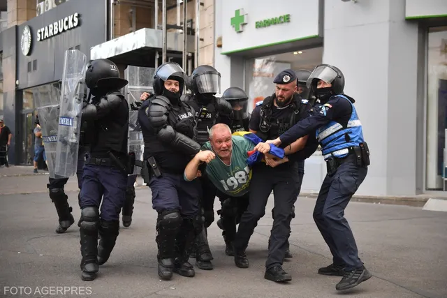 Mai mulți polițiști în echipament de protecție negru și albastru îl transportă pe un bărbat îmbrăcat într-un tricou verde pe o stradă urbană.