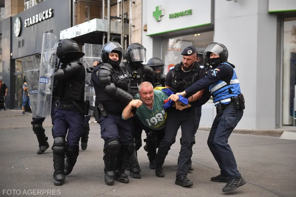 Mai mulți polițiști în echipament de protecție negru și albastru îl transportă pe un bărbat îmbrăcat într-un tricou verde pe o stradă urbană.