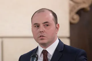 Un bărbat cu părul scurt și rare, purtând sacou închis la culoare, cămașă albă și cravată maro, stă în fața unui microfon.