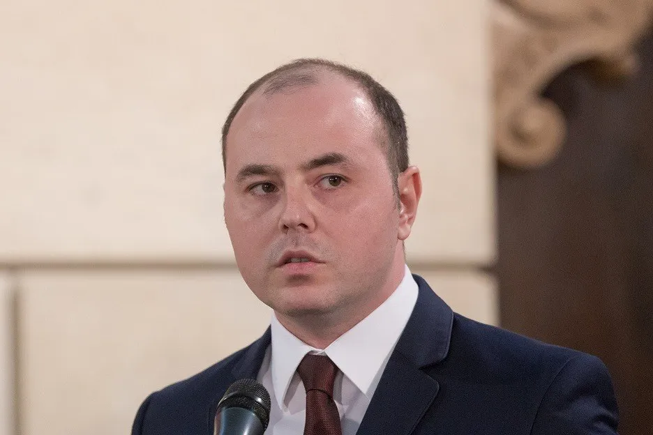 Un bărbat cu părul scurt și rare, purtând un costum închis la culoare, cămașă albă și cravată maro, stă în fața unui microfon.