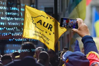 O persoană ține un telefon mobil în care se vede o mulțime, în timp ce în planul secund se ridică un steag galben cu text negru.
