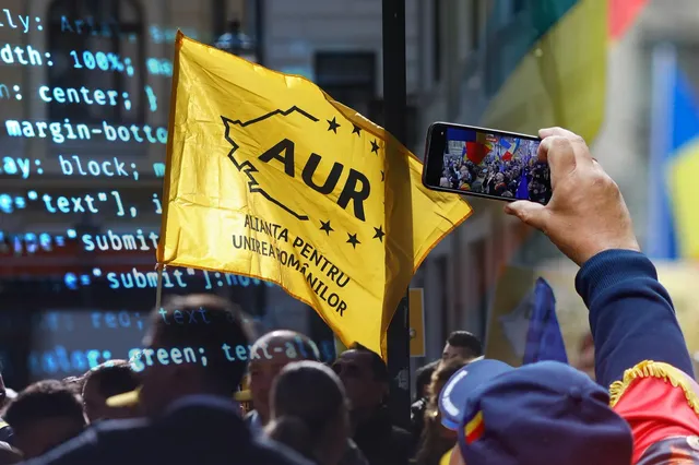 O persoană ține un telefon mobil în care se vede o mulțime, în timp ce în planul secund se ridică un steag galben cu text negru.
