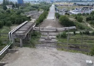 Conducte metalice suspendate trec peste câteva șine de tren și o porțiune de drum, într-un peisaj cu vegetație și clădiri îndepărtate.
