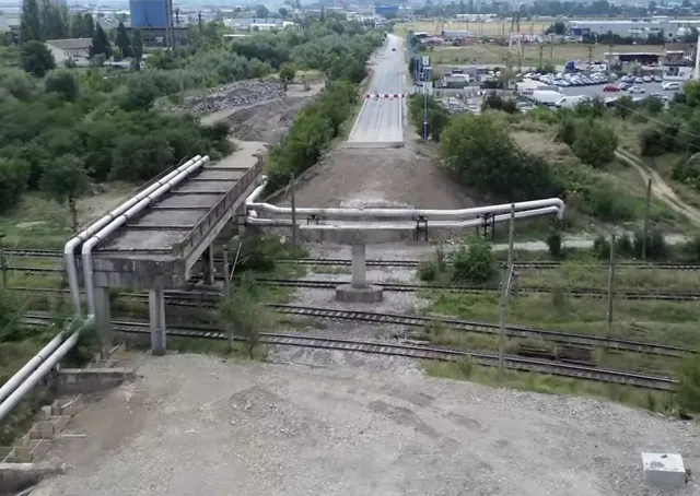 Conducte metalice suspendate trec peste câteva șine de tren și o porțiune de drum, într-un peisaj cu vegetație și clădiri îndepărtate.