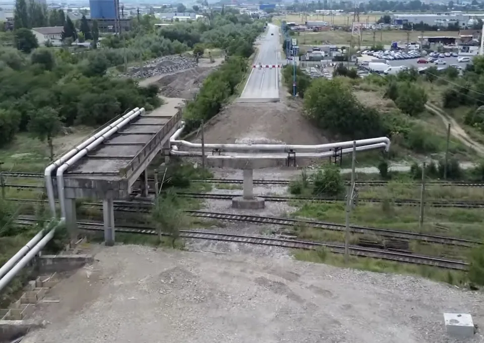 Conducte metalice suspendate trec peste câteva șine de tren și o porțiune de drum, într-un peisaj cu vegetație și clădiri îndepărtate.