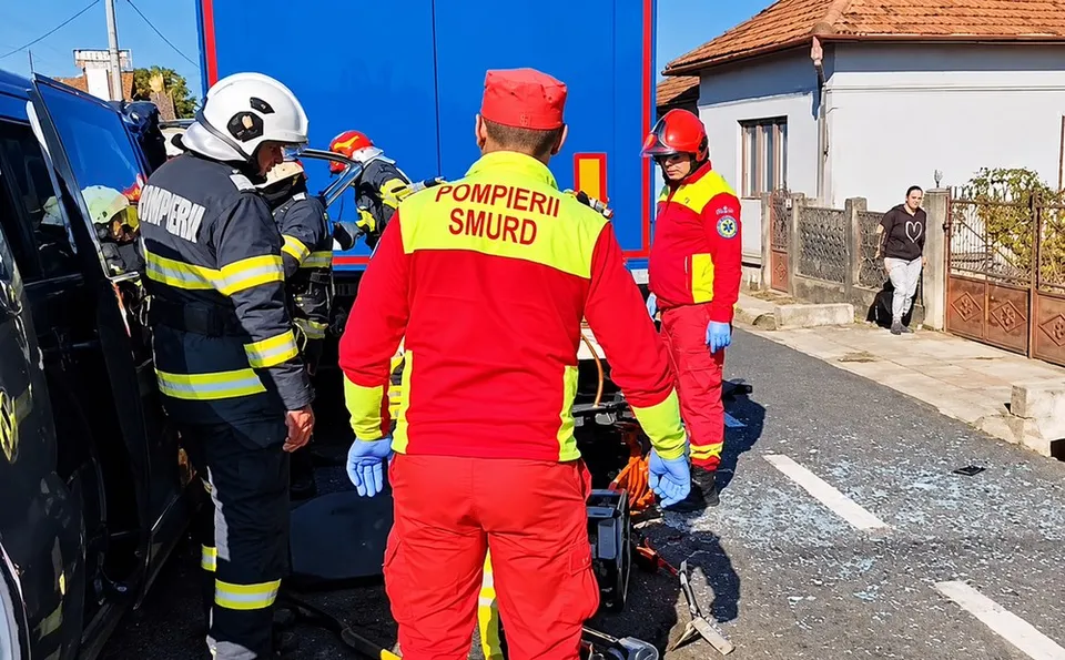 Patru persoane în uniforme de serviciu roșii și gri stau pe un drum lângă un vehicul negru și un camion albastru.