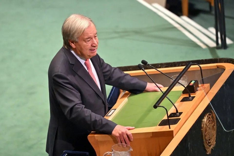 Un bărbat cu părul cărunt, îmbrăcat în costum închis și cravată roz, stă în spatele unui podium de lemn cu un panou verde.