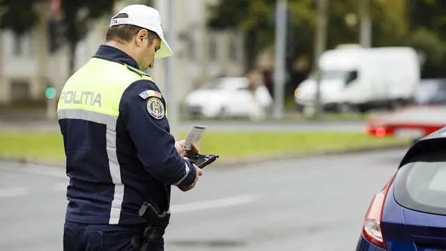 Un polițist cu vestă reflectorizantă și șapcă albă ține un document și un dispozitiv electronic lângă partea din spate a unei mașini albastre.