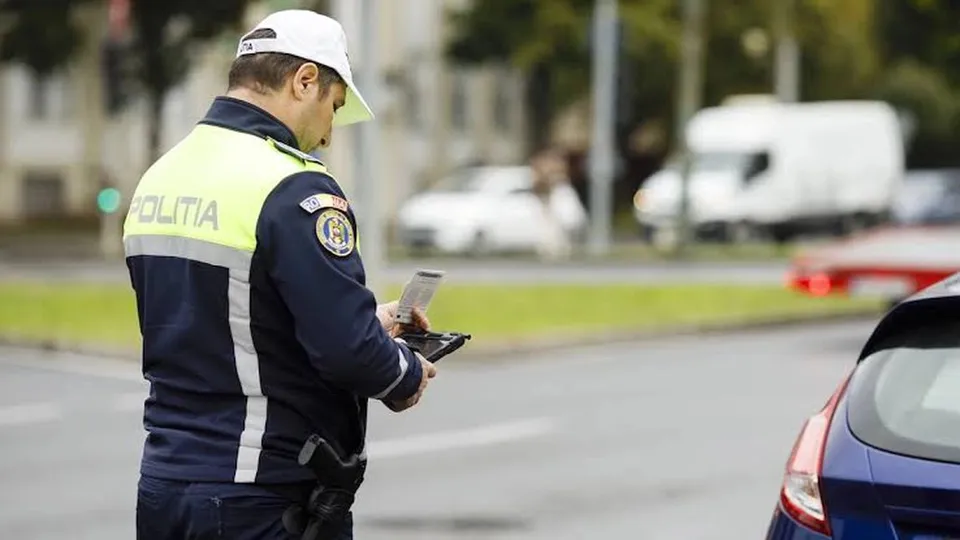 Un polițist cu vestă reflectorizantă și șapcă albă ține un document și un dispozitiv electronic lângă partea din spate a unei mașini albastre.