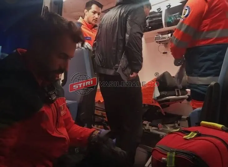 Două persoane în uniforme roșii și portocalii se află într-un vehicul lângă echipamente medicale și o geantă de transport roșie.