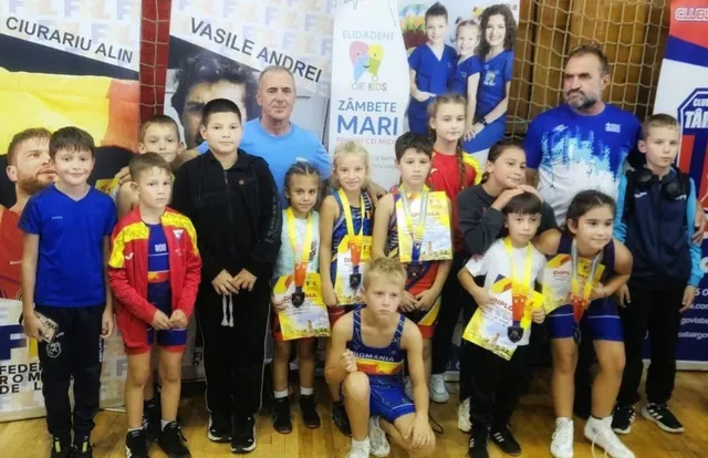 Un grup de copii și adulți stau în picioare și stau așezați, purtând medalii și echipamente sportive într-o sală de sport.