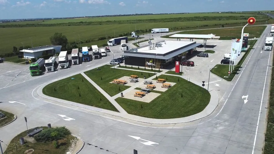 O vedere aeriană prezintă o stație de alimentare cu camioane parcate, un clădire de servicii și o zonă de gazon cu câteva bănci.