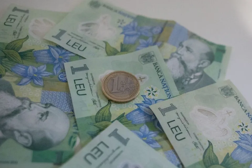 Mai multe bancnote de un leu românesc sunt împrăștiate pe o suprafață albă, având o monedă de un leu plasată deasupra uneia dintre ele.