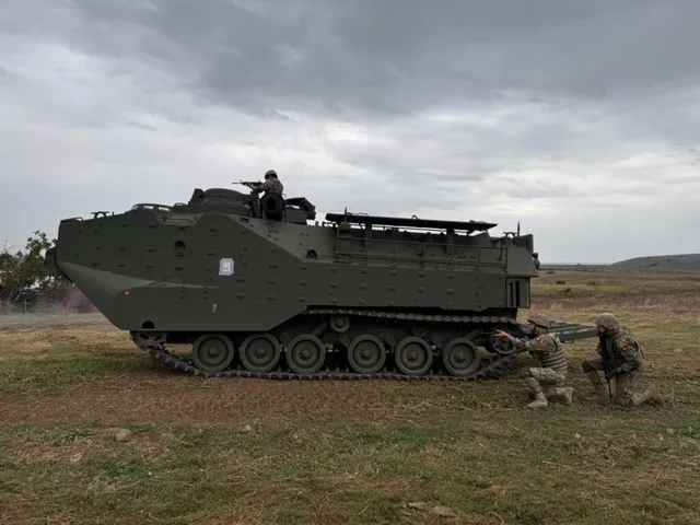 Un vehicul militar blindat pe șenile este poziționat pe un teren deschis lângă doi soldați în poziție de tragere.