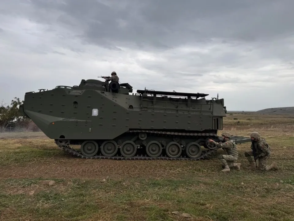 Un vehicul militar blindat pe șenile este poziționat pe un teren deschis lângă doi soldați în poziție de tragere.