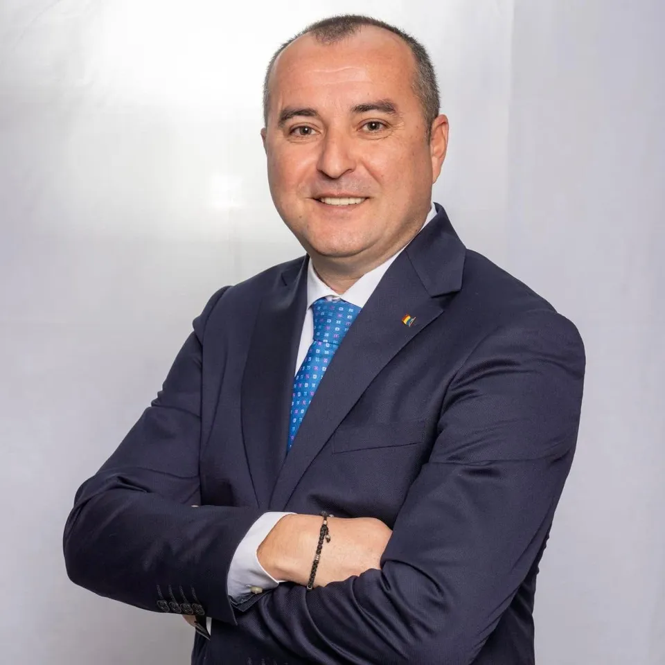 Un bărbat zâmbind poartă un costum închis la culoare, cămașă albă și o cravată albastră cu puncte albe, stând cu brațele încrucișate.