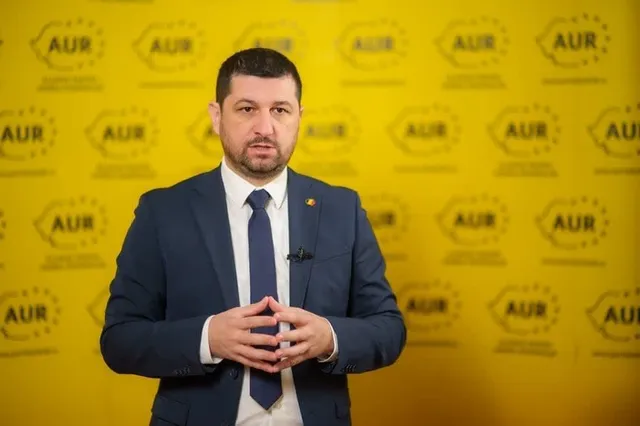 Un bărbat cu barbă scurtă, purtând sacou albastru, cămașă albă și cravată închisă, stă în fața unui fundal galben cu logouri repetitive.