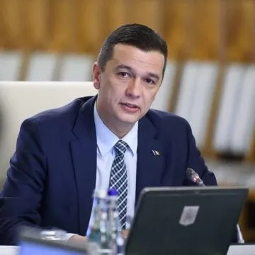 Un bărbat cu părul scurt, purtând sacou albastru, cămașă albă și cravată închisă la culoare, stă la o masă în fața unui laptop.