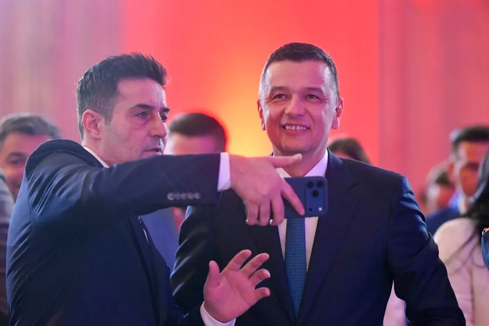 Un bărbat într-un costum închis la culoare arată spre un alt bărbat zâmbitor îmbrăcat în costum și cravată albastră.