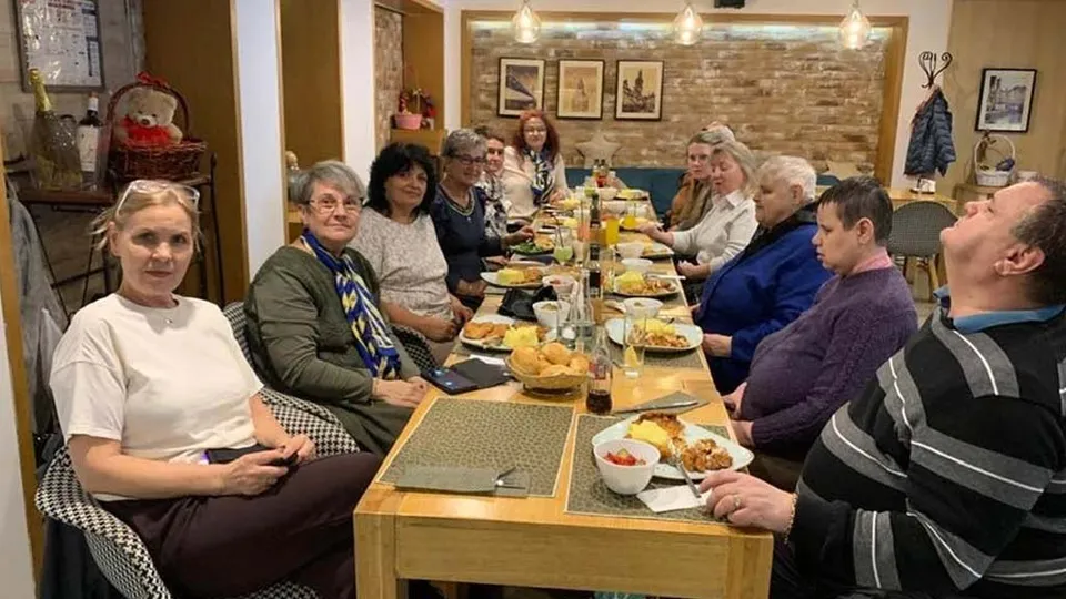 Un grup de persoane stă așezat la o masă lungă de dining într-o încăpere cu pereți din cărămidă, având farfurii și pahare în față.