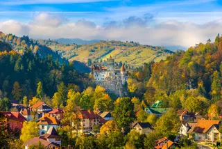 Un castel situat pe un deal stâncos este înconjurat de păduri colorate și de case cu acoperișuri în culorile toamnei sub un cer înnorat.