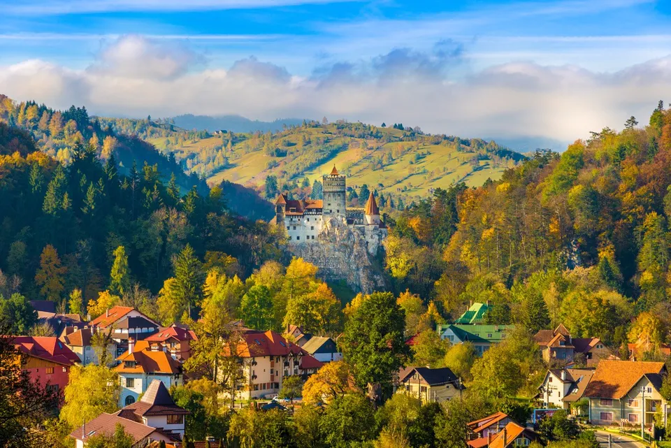 Un castel situat pe un deal stâncos este înconjurat de păduri colorate și de case cu acoperișuri în culorile toamnei sub un cer înnorat.