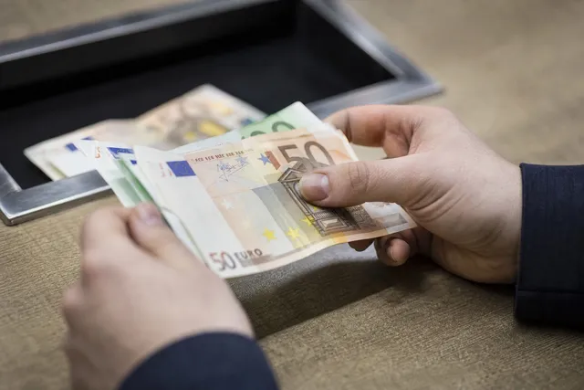 Două mâini țin și numără câteva bancnote de cincizeci de euro deasupra unei tăvițe negre.