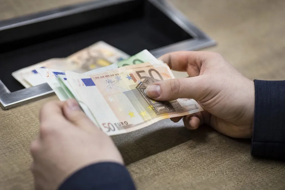 Două mâini țin și numără câteva bancnote de cincizeci de euro deasupra unei tăvițe negre.